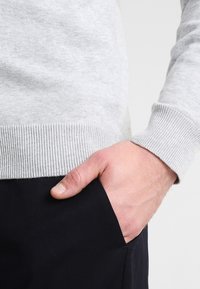 Ruka se světlou pletí odpočívající v kapse černých kalhot, oblečená v světle šedém pleteném svetru s žebrovanými manžetami a lemem.