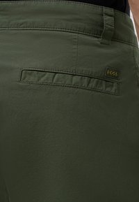 Gros plan sur un pantalon vert olive montrant une poche arrière, un passant de ceinture et une petite étiquette de la marque BOSS au-dessus de la poche.