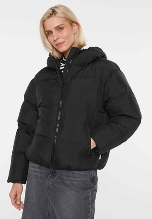Blonde Frau trägt eine schwarze gesteppte Jacke mit Kapuze, die hochgeschlossen ist, kombiniert mit dunkelgrauen Jeans und einem gestreiften Hemd darunter. Hände in den Taschen.