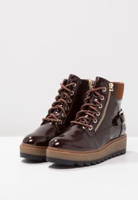 Hnědé lakované kožené kotníkové boty s tkaničkami, bočními zipy a robustní texturovanou podrážkou. Obsahují zlaté kování a kontrastní hnědou podpatek.