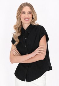 Cămașă neagră cu mâneci scurte și guler, având un model texturat, cu nasturi pe față și margine rotunjită, purtată cu pantaloni de culoare deschisă.