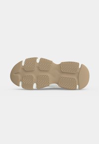 Semelle d’une chaussure beige avec des coussinets en caoutchouc texturés et rainurés pour une bonne adhérence sur un fond gris clair.