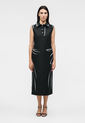 Egyenes, sötét hajú nő, aki ujjatlan fekete midi ruhát visel fehér absztrakt vonalakkal és fekete pántos magassarkú cipőben, fehér háttér előtt áll.