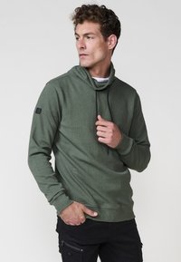 Hombre con un suéter verde texturizado de cuello alto y cordones negros, vistiendo pantalones cargo negros, mirando a su izquierda con la mano cerca del cuello y el bolsillo.