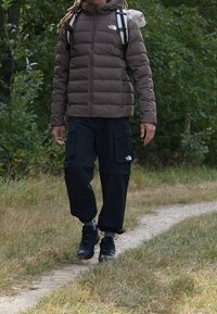 Brązowa kurtka puchowa, czarne spodnie cargo z kieszeniami, czarne buty trekkingowe, idący ścieżką otoczoną trawą i drzewami.