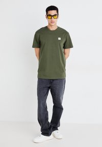 Olijfgroene t-shirt met korte mouwen en een klein logo op de borst, gecombineerd met donkergrijze spijkerbroek en witte sneakers; het model draagt gele zonnebril.