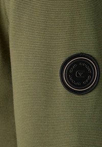 Olivgrüner strukturierter Stoff mit einem kreisförmigen schwarzen Patch, das erhabene Buchstaben aufweist. Der Stoff hat eine gerippte Oberfläche.
