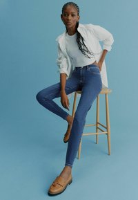Blugi skinny bleumarin, tricou alb ajustat, jachetă deschisă la culoare și mocasini maro. Stând pe un taburet din lemn, pe un fundal albastru.