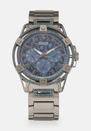 Höyhenhopeinen ruostumaton teräsranneke, jossa on sininen analoginen kellotaulu, kolme alinäyttöä ja kristallikoristeinen kehys, jossa on teksturoitu kuvio.