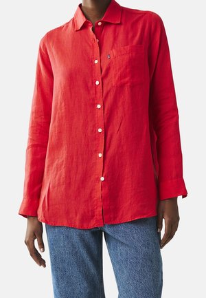 Person wearing a long-sleeve red button-up shirt with a chest pocket paired with blue denim jeans, standing against a plain background.