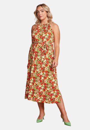 Kobieta ubrana w bezrękawnikową sukienkę na szyję w kwiaty w kolorach czerwonym, zielonym i kremowym, dopasowaną w talii, do tego zielone szpilki.