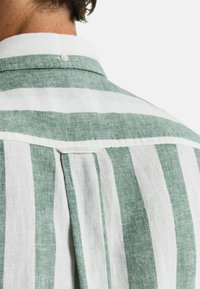 Camisa de lino a rayas verdes y blancas con cuello abotonado y un plisado en la parte posterior, que destaca su diseño relajado.