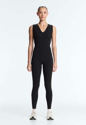 Stehende Person, die einen figurbetonten schwarzen ärmellosen Overall und weiße Sneakers vor einem schlichten, hellen Hintergrund trägt.