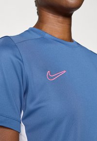 Μπλε αθλητικό μπλουζάκι με κοντό μανίκι, διακοσμημένο με το ροζ κεντημένο λογότυπο της Nike στο στήθος. Κατασκευασμένο από υφαντό, υλικό που απορροφά την υγρασία.