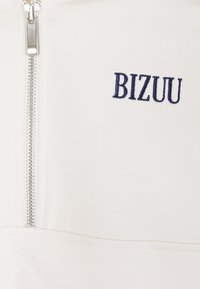 Crème ritsweater met marineblauw geborduurd "BIZUU" logo. Heeft een zilveren rits, een gladde textuur en een getailleerd ontwerp.