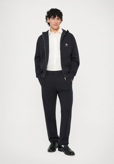 Sudadera de un oscuro azul marino con cremallera plateada y logo bordado, combinada con pantalones de chándal a juego, llevada sobre una camisa blanca con botones y zapatos negros.