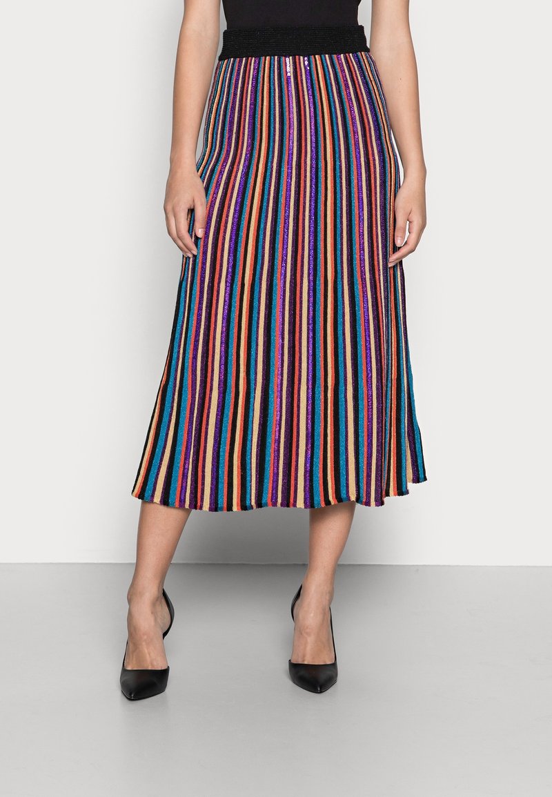 Femme portant une jupe midi plissée avec des rayures verticales multicolores et des talons hauts noirs, se tenant devant un fond clair uni.