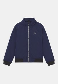 Námořnická modrá bunda na zip s vysokým límcem, černými žebrovanými manžetami a lemem, s malým bílým logem CK a vzorovaným detailem na zipu.