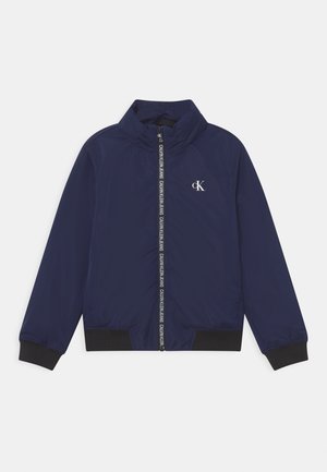 Marineblauwe zip-up jack met hoge kraag, zwarte ribbel manchetten en zoom, met een klein wit CK-logo en een patroon op de rits.
