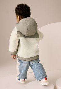 Veste beige clair et grise doublée en polaire avec capuche, portée avec un jean bleu ample et des baskets blanches avec des accents rouges et bleus.