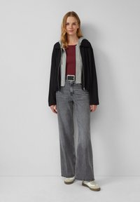 Veste zippée noire superposée sur un sweat à capuche gris, associée à un haut bordeaux, un pantalon gris large et des baskets beiges avec des accents blancs.