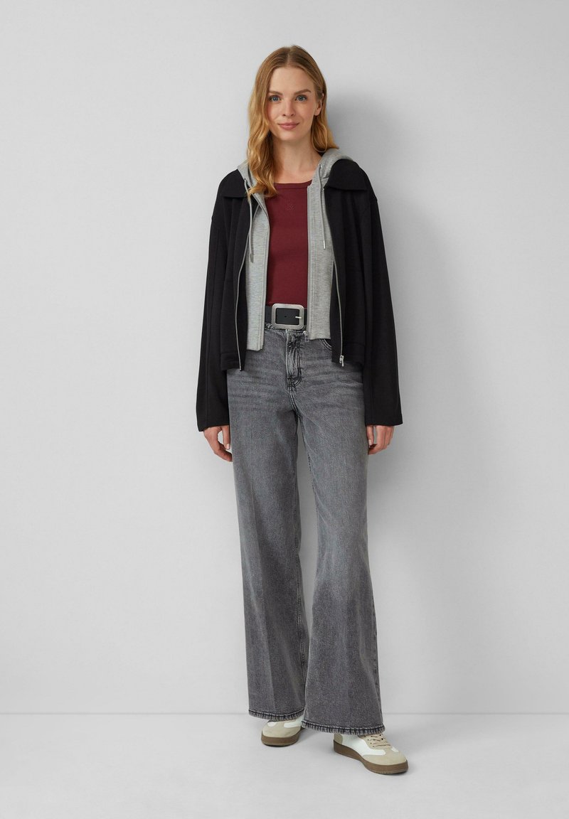 Veste zippée noire superposée sur un sweat à capuche gris, associée à un haut bordeaux, un pantalon gris large et des baskets beiges avec des accents blancs.