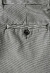 Tissu texturé gris avec une petite poche avant et un bouton noir. Le tissu présente un motif tissé subtil et une finition lisse.