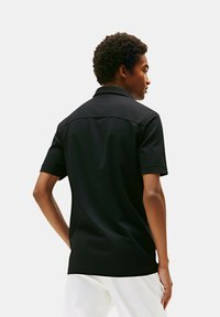 Polo shirt nero a maniche corte con colletto, caratterizzato da una texture liscia e un design aderente. La vista posteriore mostra le cuciture sulle spalle e linee pulite.