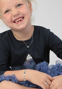 Zilveren sieraden bij een kind: ketting met een harthanger, armband met een hartaccent, gekleed in een marineblauwe top met een paillettenster en een blauwe gerende rok.
