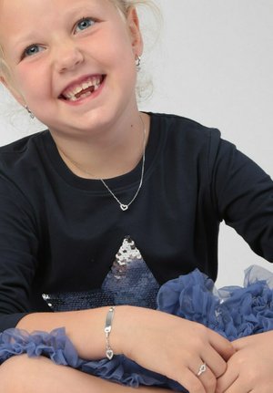 Zilveren sieraden bij een kind: ketting met een harthanger, armband met een hartaccent, gekleed in een marineblauwe top met een paillettenster en een blauwe gerende rok.