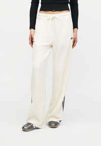 Γυναίκα που φοράει άνετο λευκό παντελόνι Adidas με μαύρες ρίγες και μαύρο κοντό μακρυμάνικο τοπ, στέκεται με τα χέρια χαλαρά στα πλάγια.