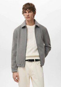 Giacca in pile grigia con colletto, chiusura con cerniera e tasche laterali, indossata sopra un maglione crema a trecce e pantaloni chiari.