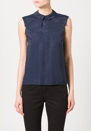 Marineblå ærmeløs bluse med rund krave, blød glans, plisseringsdetaljer ved skuldrene og en afslappet pasform, parret med mørke jeans.