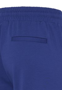 Aizmugures skats uz zila elastīga vidukļa biksēm ar vienu horizontālu aizmugures kabatu bez aizdares.