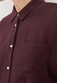 Camicia bordeaux con bottoni, realizzata in tessuto liscio, caratterizzata da un colletto classico, bottoni bianchi e una tasca sul petto. Design semplice e pulito.