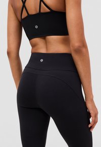 Donna che indossa un reggiseno sportivo nero Lululemon con spalline e leggings a vita alta abbinati, mostrata di spalle su uno sfondo neutro.