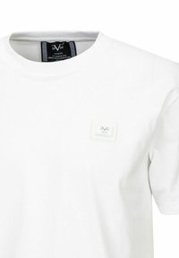 Witte katoenen t-shirt met een ronde hals, voorzien van een kleine rechthoekige logo patch in groen en blauw op de borst. Glad textuur, korte mouwen.