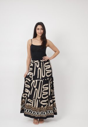 Mujer con pelo largo y oscuro, vistiendo una blusa negra sin mangas y una falda larga negra con patrones abstractos blancos, de pie frente a un fondo blanco.