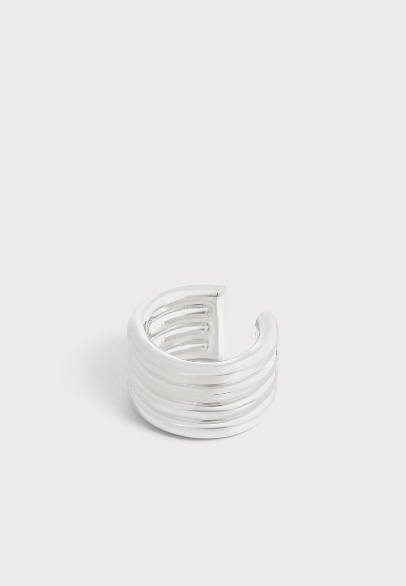 Une bague ouverte en argent poli, de style manchette, avec plusieurs reliefs horizontaux, sur un fond uni clair.