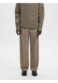 Brūns adīts džemperis ar ribotām aprocēm, kombinēts ar bēšiem plaša piegriezuma biksēm un melnām loaferēm, uzsverot faktūru un pielāgotu dizainu.