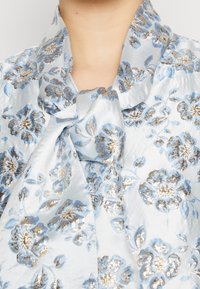 Vaaleansininen kukkakuvioinen paita satiinipinnalla, jossa on metallinhohtoisia kultaisia yksityiskohtia ja solmittu nauha kauluksessa, yksityiskohtana tekstuuri.