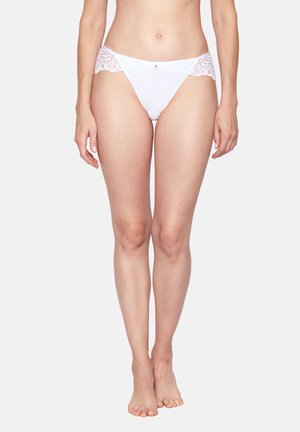 Frau steht barfuß in weißer, mit Spitze gesäumter Bikini-Unterwäsche vor einem schlichten hellen Hintergrund.