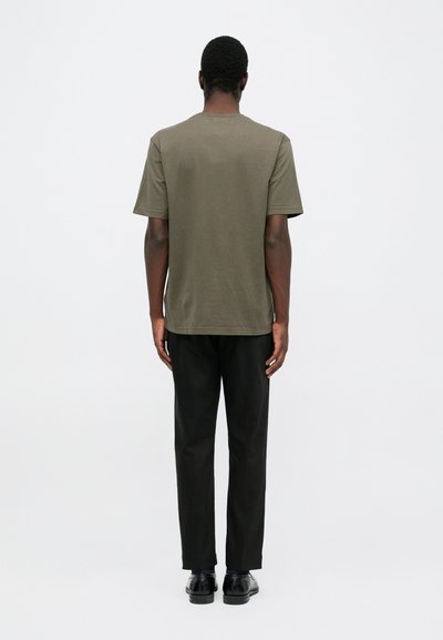 T-shirt kaki a maniche corte con colletto rotondo, abbinato a pantaloni neri e scarpe nere, visto da dietro su uno sfondo bianco.
