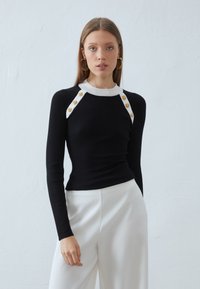 Musthave mustriba pikkade varrukatega topp valge kaelusega ja õlavarre aksessuaaridega. Omab kuldseid nuppe õlavarre õmblustel. Kokku sobitatud valgete pükstega.