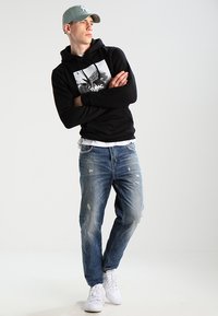 Schwarze Kapuzenjacke mit Grafikdruck, blaue ausgebleichte Jeans und weiße Sneaker; trägt eine hellgraue Mütze, Arme verschränkt, neutraler Hintergrund.