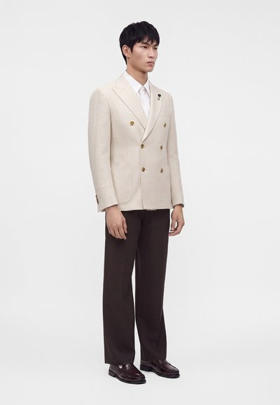 Homme portant un blazer croisé beige, chemise blanche, pantalon marron foncé et chaussures habillées marron foncé, debout devant un fond uni.