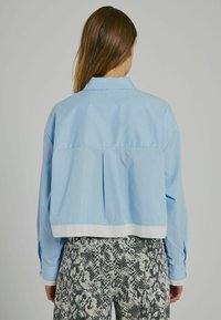 Camicetta azzurra chiara corta con dettagli a pieghe sul retro e orlo bianco a contrasto. Tessuto morbido con maniche lunghe e polsini a bottoni.