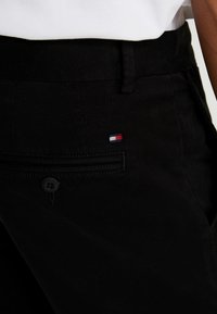 Černé kalhoty s kapsou na zip, zapínáním na knoflík a malou červenou, bílou a modrou nášivkou na zadní kapse. Hladká textura.