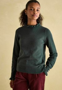 Dunkelgrüner Pulli mit einem Rüschenausschnitt, geripptem Saum und Knopfdetails an den Ärmeln. Der Stoff wirkt weich und strukturiert. Kombiniert mit burgunderfarbenen Hosen.