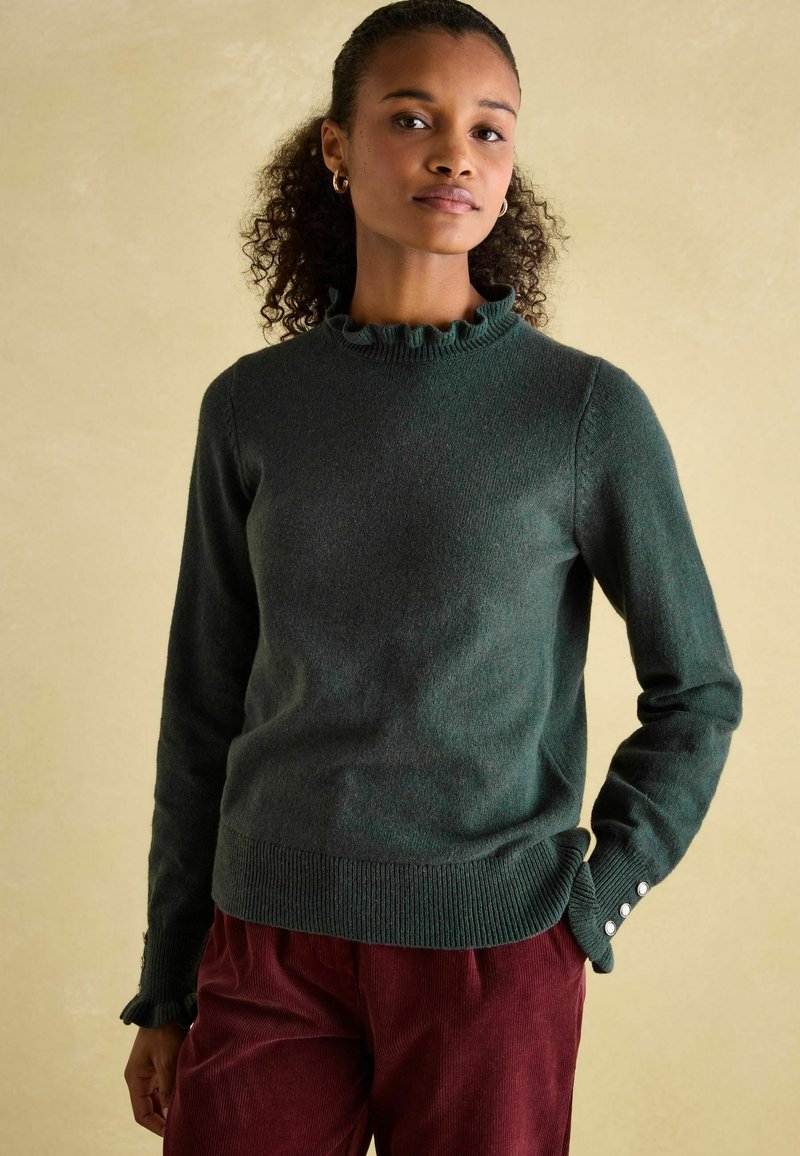 Dunkelgrüner Pulli mit einem Rüschenausschnitt, geripptem Saum und Knopfdetails an den Ärmeln. Der Stoff wirkt weich und strukturiert. Kombiniert mit burgunderfarbenen Hosen.
